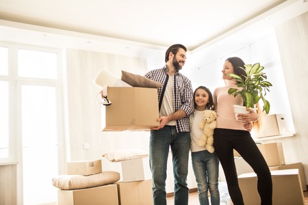 A picture of good family standing together in room. Parents are looking to each other and smiling. Man is holding a box in his hands while the woman has got a plant. There is a small girl between them.の写真素材