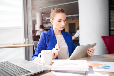 Smart and good-looking woman is sitting on the table and looking to the tablet. She is studying. Girl is very concentrated on subject.の写真素材