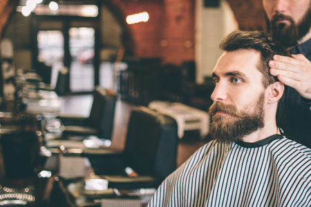 Bearded guy is sitting in salon. His hairstylist is modelling his hair. Guy is looking straight forward.の写真素材