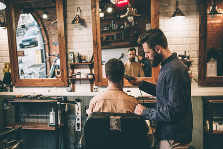 Nice pictur is a barber shop. Two guys are there. One of them is cutting hair with electrical razer while the other one is sitting in chair.の写真素材