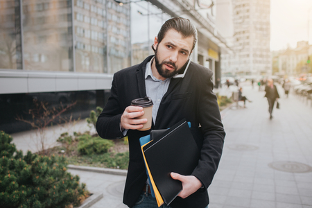 Busy man is in a hurry, he does not have time, he is going to talk on the phone on the go. Businessman doing multiple tasks on the hood of the car. Multitasking business person is drinking cofeeの写真素材