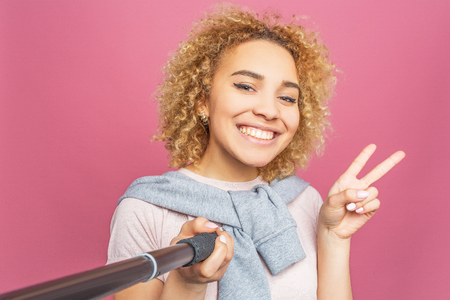 Smiling girl is taking a selfie. She uses a selfie sticj for that. Young woman is posing and showing the piece symbol to camera. Isolated on pink backgroundの写真素材