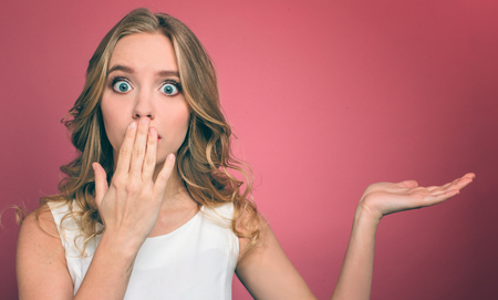 Girl with blond hair and big blue eyes is amazed. She is covering her mouth with hand and posing with the other hand. Isolated on pink background.の写真素材