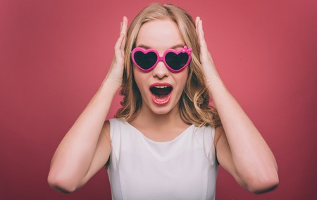 Amazed and attractive girl wears sunglasses. She is looking straight forward. Female person is holding her hands on the head. Isolated on pink background.の写真素材