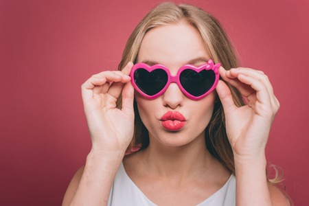 Adorable girl in glasses with pink edge is looking straight forward. he is holding glasses on its edge close to the head. Yng woman is making a kiss shape from her lips. Isolated on pink background.の写真素材