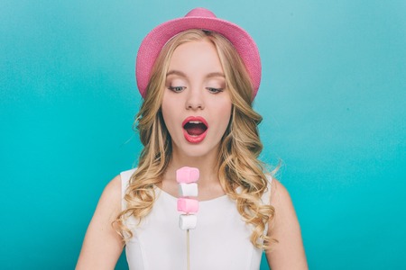 Amazed and excited girl is looking down to pieces of marshmallow that are on the stick. She is going to eat it. Girl has opened her mouth to eat it. Isolated on blue background.の写真素材