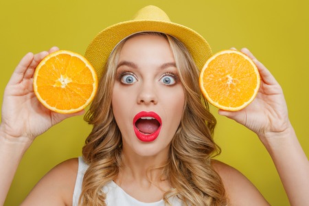 Amazed and excited young woman wears yellow hat. Also she is showing two pieces of juicy orange to...