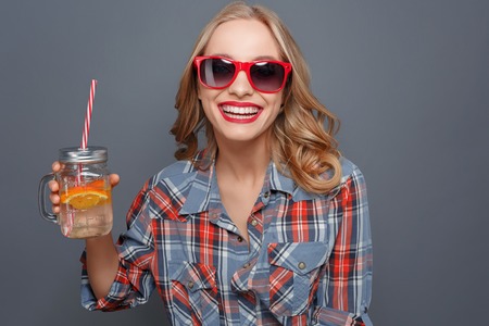 Awesome and nice girl is holding a cup with some juice and a piece of orange in it. She is laughing. Girl wears dark glasses with red edge. Isolated on grey background.の写真素材