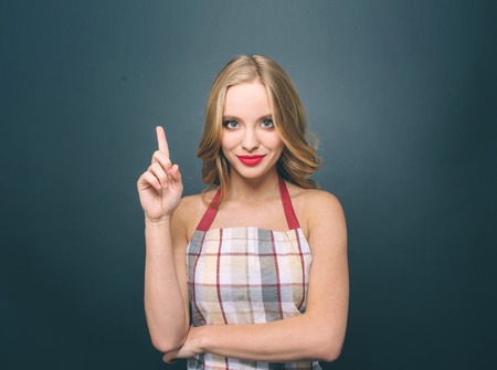 Sexy blonde girl is standing and pointing up. She looks fantastic. Also girl wears apron. Isolated on blue background.の写真素材