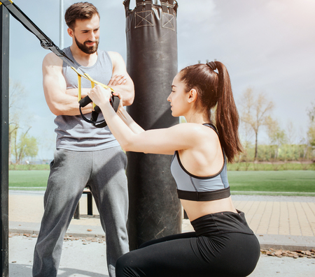 Vertical picture of a slim girl doing squats. She is holding the loops to keep balance. Young man is...