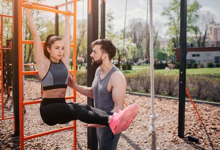 Beautiful couple is exercising outside in park. Girl holds horizontal bar and keeping her legs in a...