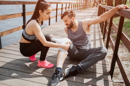 Man is sitting on the bridge and holding the wood panel with the hand. He has a pain in his knee. Girl is sitting besides him and helping to wrap his knee up.の写真素材