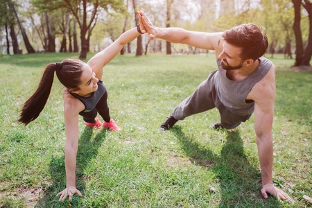 Slim and well-built people are exercising on grass in park. They are staying in plank position and giving high-five to each other. Also young man and woman are looking to each other and smiling.の写真素材