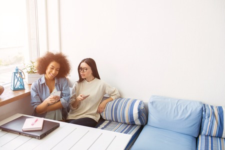 Nice picture of students sitting close to each other on sofa. THey are holding phones in their hands. Afro american girl is smiling while her friends is not happy and wondering. They have a break now.の写真素材