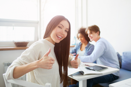 Nice picture of asian girl showing big thumb up. She looks happy. Also there are another people sitting at the table. They are studying.の写真素材
