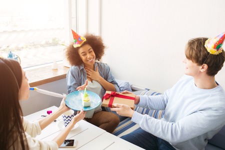 Happy friends are celebrating birthday. They have funny hats on their heads. Guy is holding a...
