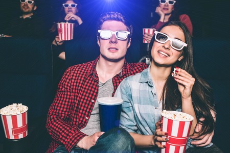 Two young people are sitting close to each other. They have special glasses on their faces to watch movie. Girl is holding basket with popcorn and one piece of it in her hands. Man has a cup of coke.の写真素材