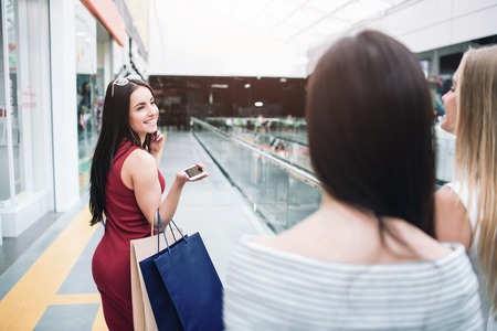 Cheerful and beautiful girl in red dress is looking back to her friends and smiling. Also she holds two bags and phone in her hands. She looks happy.の写真素材