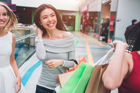 Cut view of positive and happy girls walking together. Asian girl is holding her hair and smiling. Her blondy friend is walking besides her. Also there is a hand of girl in red dress holding bags.の写真素材