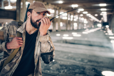 Calm and quiet fighter in uniform is speaking into portable radio. There is a rifle that is hanging over right shoulder of fighter. Man is holding its belt with right hand.の写真素材