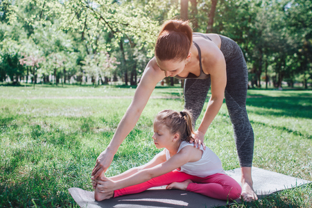A picture of woman standing upon her daughter and teaching her to stretch. Small girl is reacing her foot with her hands and keeping other foot close to left leg. Yoga and Pilates Concept.の写真素材