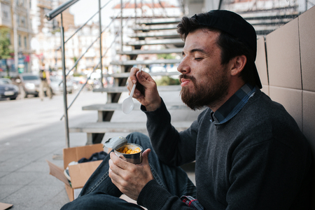 Dirty man is eating corn from can and enjoying it. He is keeping his eyes closed and smiling. Guy is sitting on the ground.の写真素材