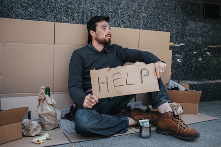 Dirty man is sitting on the ground and holding a help cardboard in hands. He is looking to the side. There are lots of stuff near him. Also there are a cup with money in front of him.の写真素材