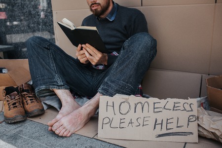 Smart homeless man is sitting on cardboard and reading a book. He is very concentrated on that....