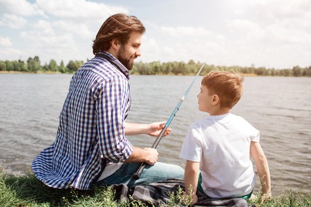 Father and son are sitting together on grass and looking at each other. Guy is holding fishing rod...