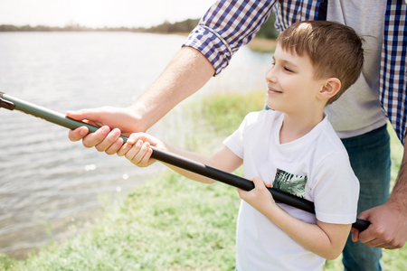 A picture of guy helping his son to hold fishing rod in a right way. He is holding it with one hand while boy is doing that with two hands. Small man seems like happy boy.の写真素材