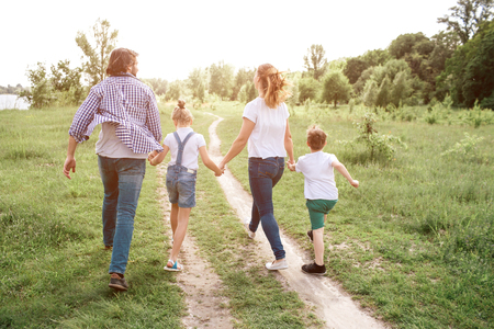 Happy family is walking down the road in meadow. Parents are holding their kids by hands. They are jumping and enjoying the moment.の写真素材