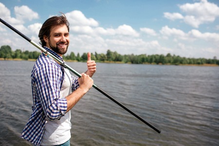 Happy guy is standing near river and looking on camera. He is smiling. Guy is holding fishing rod with right hand on right shoulder. Also man is pointing up. He looks happy.の写真素材