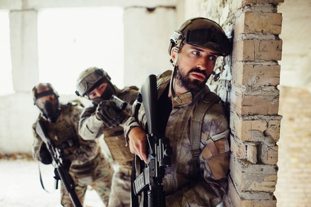 Another picture of man in uniform standing behind wall and waiting. There are tow guardians behind bearded man. They are covering him. Man in front is holding his rifle down.の写真素材