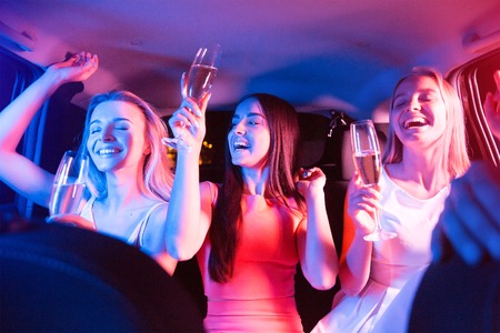 Beautiful girls are parting in car. They are having a lot of fun. They are moving with their hands, laughing and keeping eyes closed. Blonde girl and brunette girl are holding glasses of champagne.の写真素材