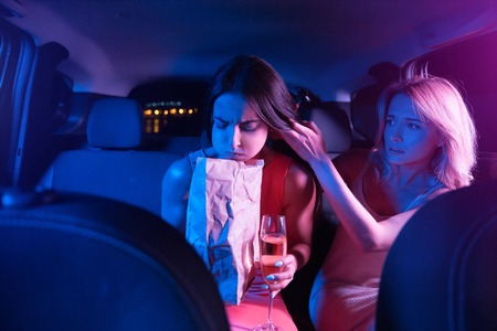 Brunette girl is sick. She is vomiting. Young woman has a paper bag in her hands. She is looking down. Blonde girl is hepling her and holding her hair. She is worried.の写真素材