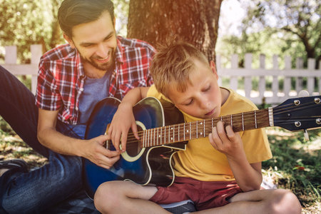 Talented boy is looking on strings and playing guitar. His father is sitting besides him and helping him. Adult is smiling.の写真素材