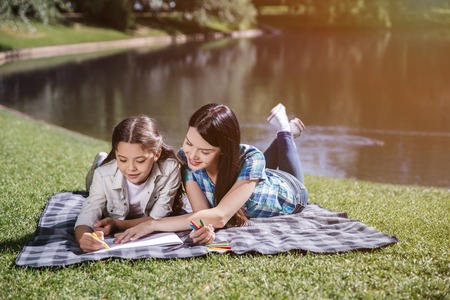 Happy mom is lying on blanket with her daughter. She holds her hand on piece of paper. Girl is drawing with yellow marker. They are smiling and looking down.の写真素材