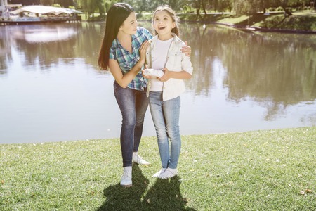 Mother is standing outside in park with her daughter. She is embracing and looking at her daughter. Girl is looking up. She is standing besides her mother. Girl is holding control panel in hands.の写真素材