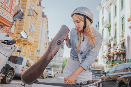 Smart and good-looking girl is looking under motorcycle seat. She is looking for something. Girl is standing outside on street.の写真素材