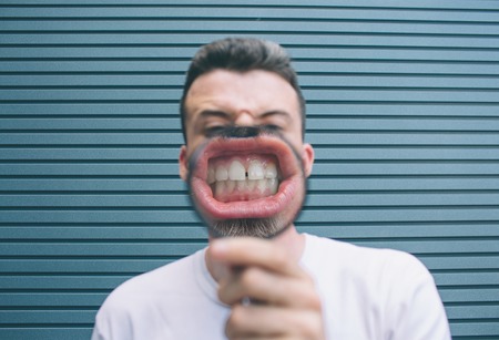Brunette in white shirt is standing at wall and posing. He is holding loup in hand and showing his teeth on camera. Guy has big teeth. Isolated on striped and blue background.の写真素材
