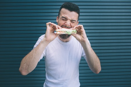 A picture of bearded man biting a piece of sandwich. He is holdng it with both hands. Guy is keeping eyes closed. Isolated on striped and blue background.の写真素材