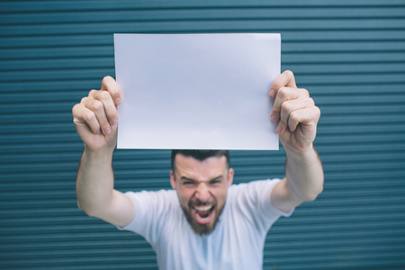 Strong guy is standing and screaming. He is yelling on camera. Man is holding blank page with both hands. He is showing it on camera. Isolated on striped and blue bsckground.の写真素材