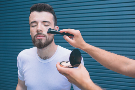 Beautiful man is standing with eyes closed. Somebody is putting make up on his face. Guy is waiting patiently. Isolated on striped and blue background.の写真素材