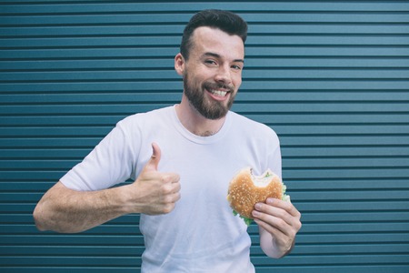 A picture of guy holding burger and showing his big thumb up. He is smiling and looking on camera....