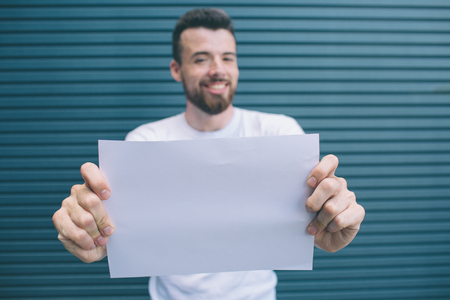Nice and good-looking man is looking on camera. He is holding blank piece of paper with both hands. He is showing it to camera. Isolated on striped and blue background.の写真素材