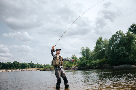 Fisherman stands in shallow and holding fly rod up in one hand and part of spoon in the other one. Man is looking up. He is calm and concentrated.の写真素材