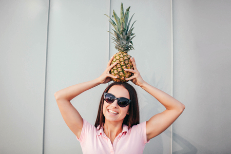 Positive girl in sunglasses stands and holds pineapple on her head. She keeps it in balance with hands. Girl looks on camea and smiles. Isolated on white and striped background.の写真素材