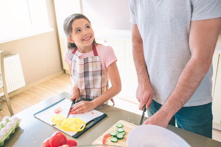 A oicture of girl cutting tomato and looking at father. She smiles. Dad stands close to her and working. Cut view.の写真素材