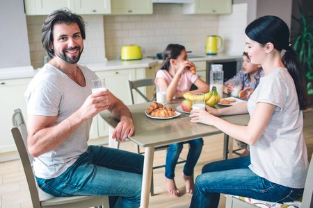 Happy man is sitting with his family at table. He holds cup of milk. Other members of family are eating food and talking with each other.の写真素材