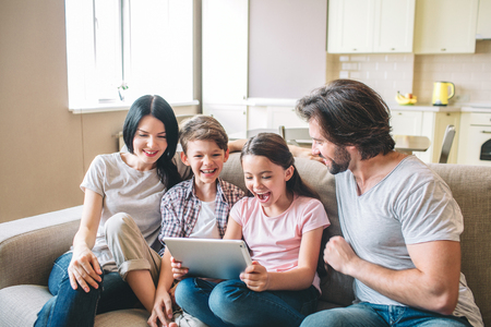 Happy family is sitting on sofa. Girl holds tablet in hands. She is excited and amazed. All members...